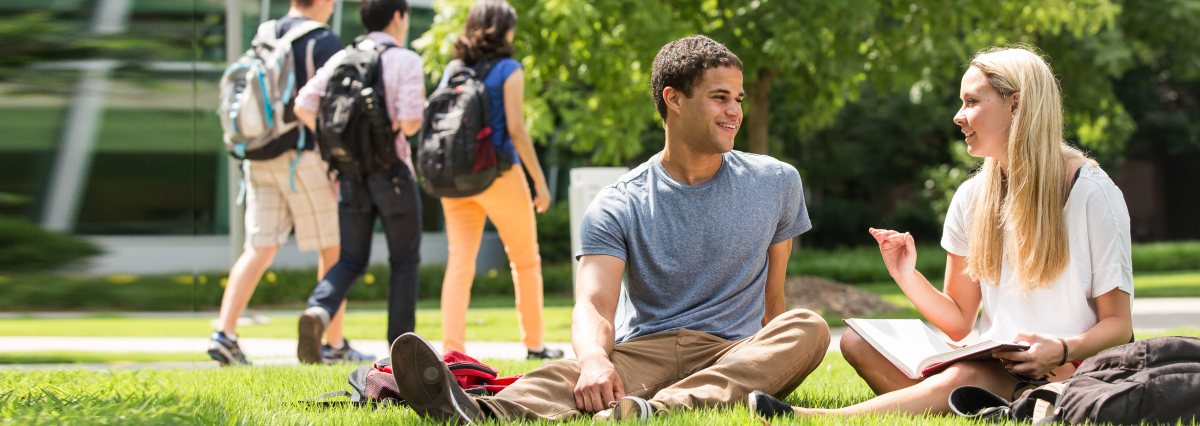 A black young man and a white young woman talk while sitting on a grass lawn.